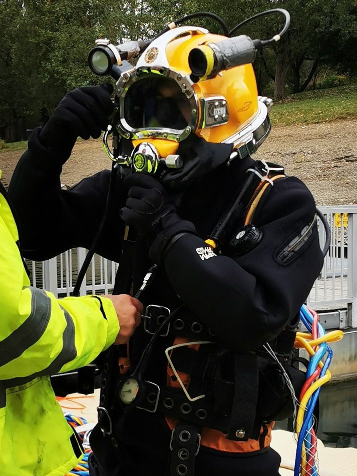 Taucher in Schutzanzug und Helm bereitet sich auf Tauchgang vor, unterstützt von einem Kollegen