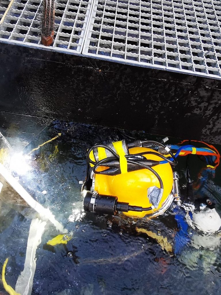 Taucher mit gelbem Helm schweißt unter Wasser an einem Metallgerüst